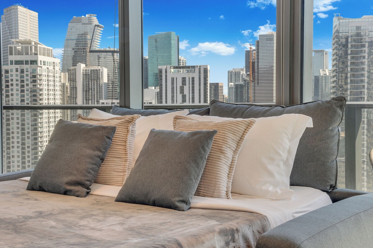 A bed with multiple pillows and a view of a cityscape through large windows.