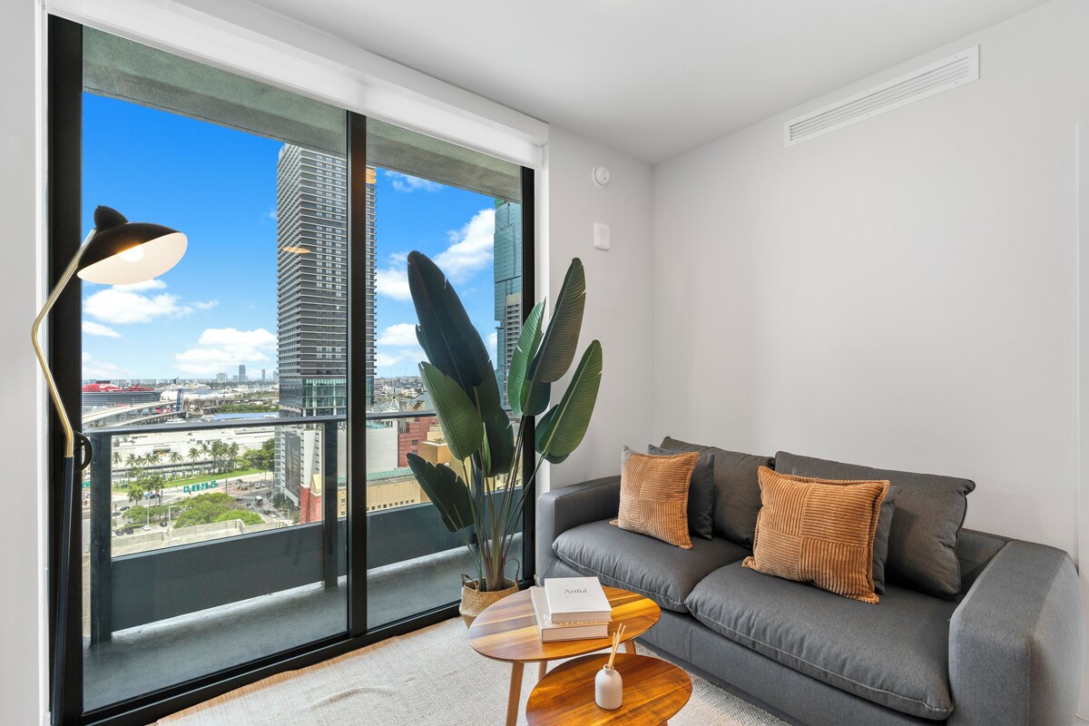 A modern living room with a grey sofa, a small wooden coffee table, a large potted plant, and a view of a cityscape through floor-to-ceiling windows.