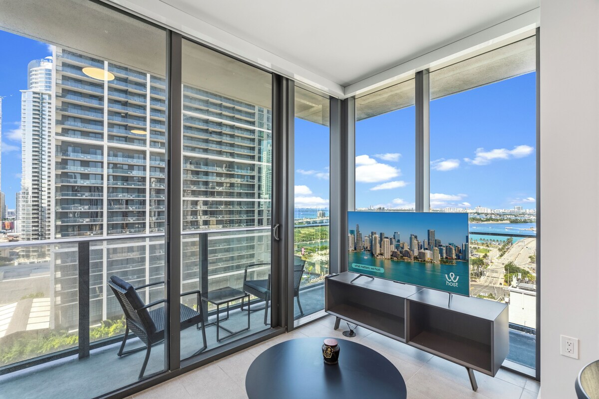 A modern room with large windows, a flat-screen TV, a round table, and a bench.