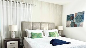 Una cama bien hecha con colcha blanca, dos almohadas y una toalla azul. Hay una mesita de noche con una lámpara y un cajón. La habitación tiene una cortina y una pared con un cuadro floral.