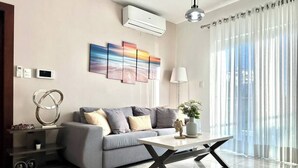 Una sala de estar moderna con un sofá gris, una mesa de centro, un aire acondicionado instalado en la pared y un ventanal con cortinas de gasa.