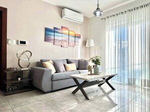 A modern living room with a grey sofa, a coffee table, a wall-mounted air conditioner, and a large window with sheer curtains. - Soha II Deluxe Suites (Santiago de los Caballeros)