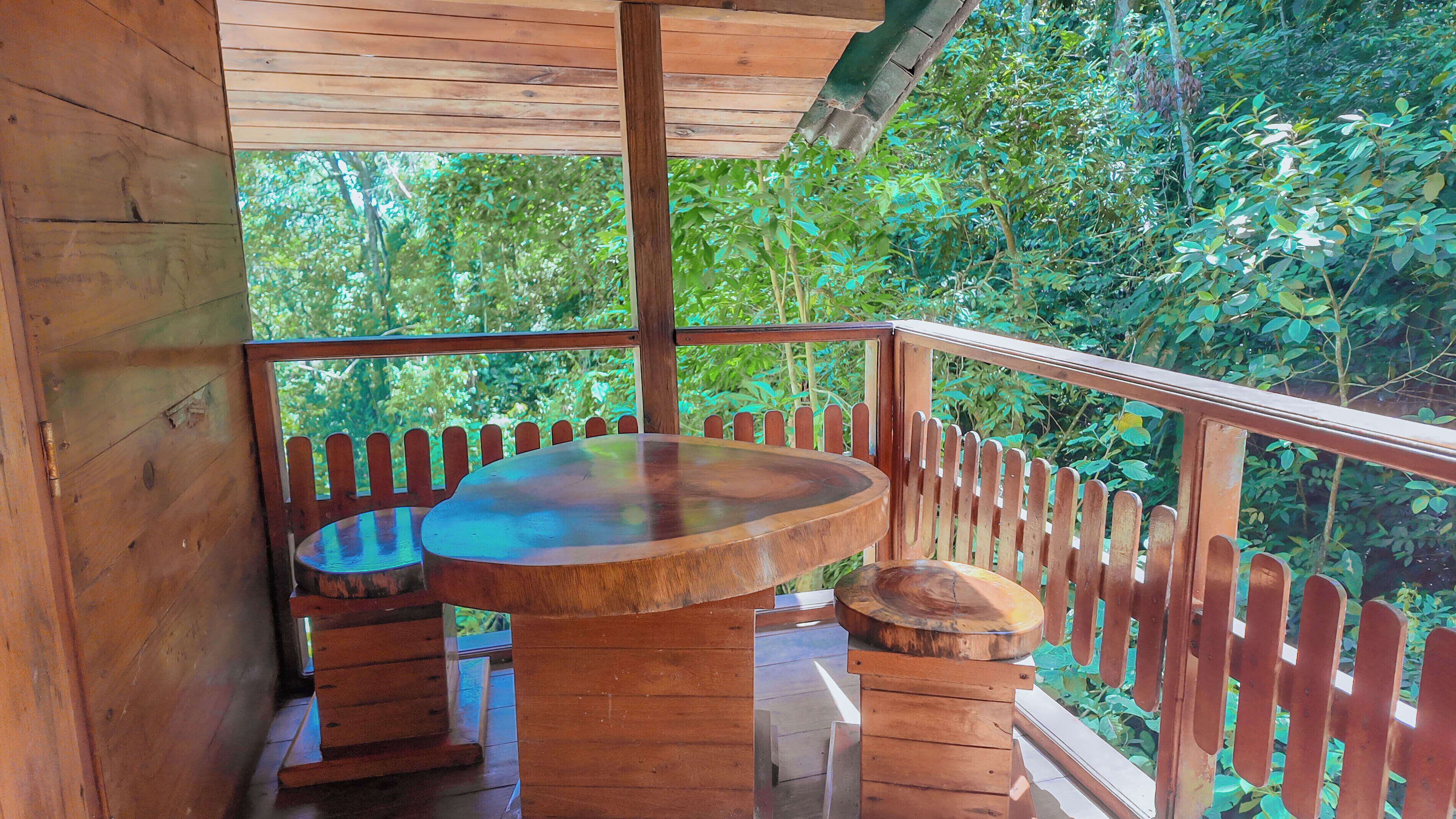A wooden deck with a round table and benches, surrounded by a wooden fence, overlooking a lush green forest.