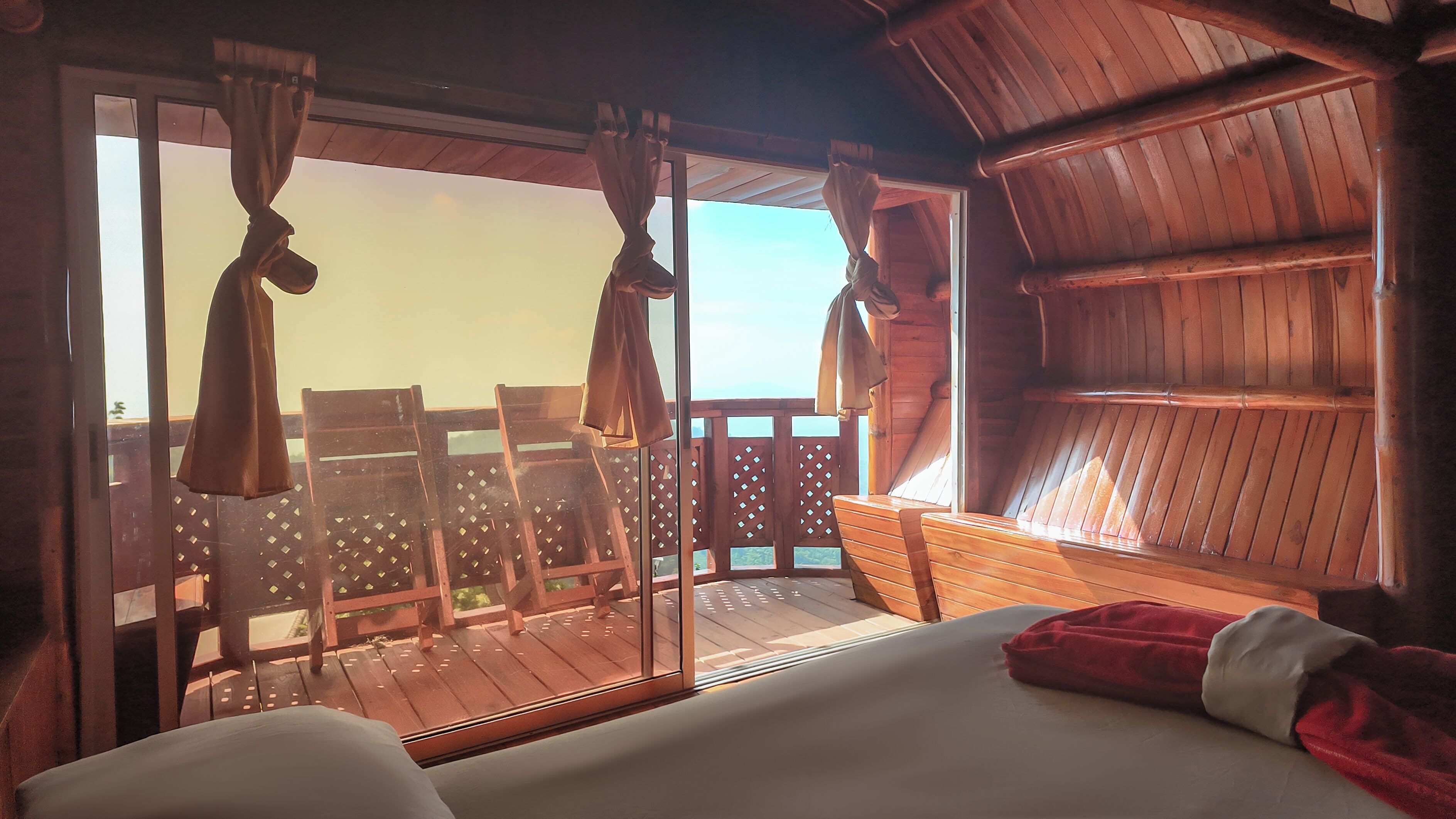 Une chambre dotée d’un grand lit, d’un plafond en bois et offrant une vue sur une terrasse extérieure aménagée de chaises, accessible par une porte vitrée.
