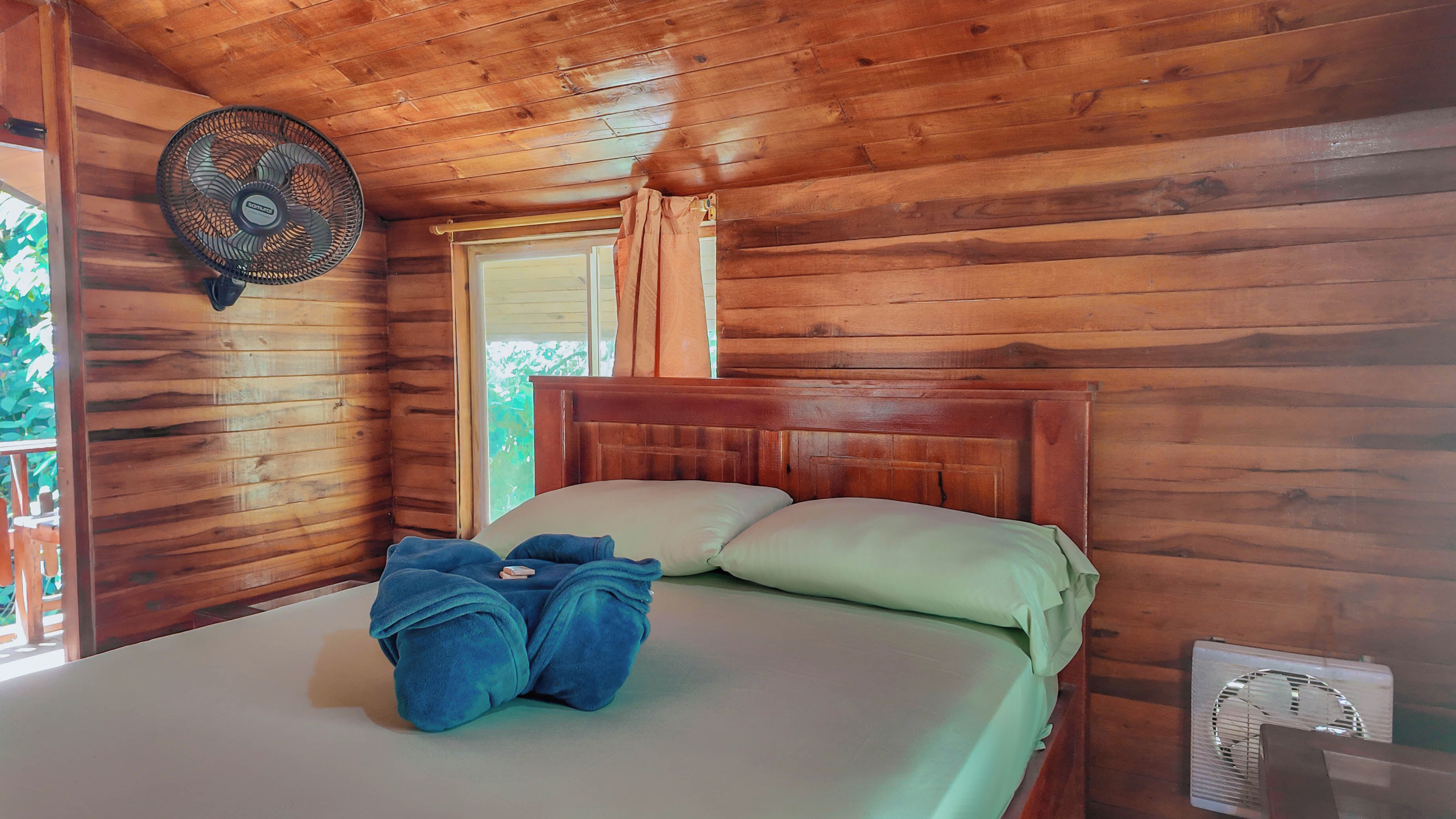 A wooden cabin room with a bed, a fan, and a window with curtains.