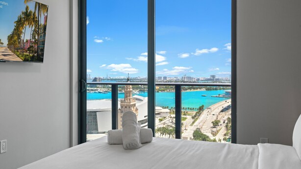 A hotel room with a large window offering a view of a coastal cityscape, a bed with white linens, and a painting on the wall. - 501 Building - Close to Kaseya Center - We Host (Miami)