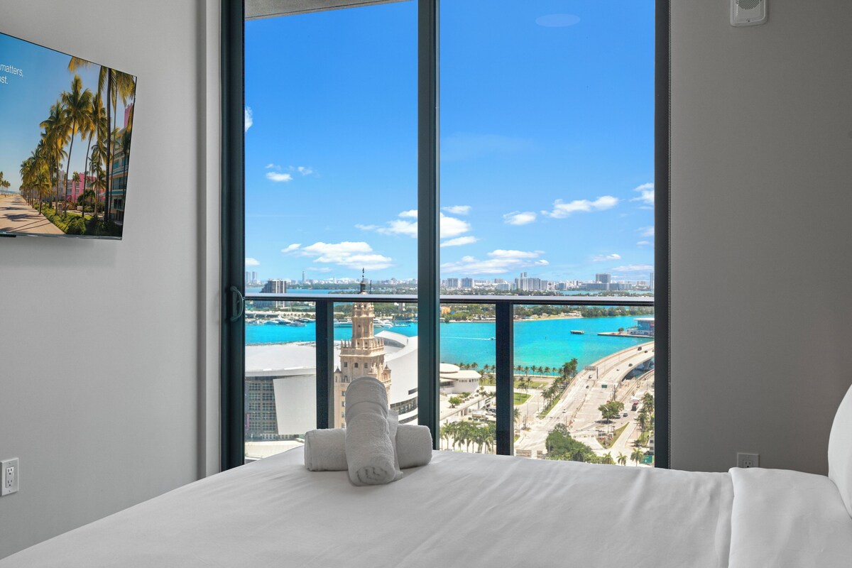 Ein Hotelzimmer mit großem Fenster und Blick auf eine Küstenstadt, einem Bett mit weißen Leinen und einem Gemälde an der Wand.