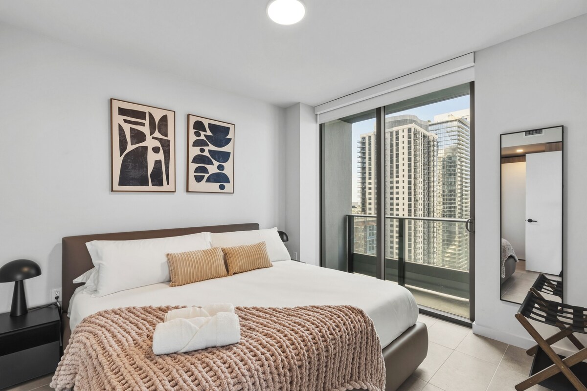 Ein modernes Hotelzimmer mit einem großen Bett, Blick auf hohe Gebäude und einem Balkon.