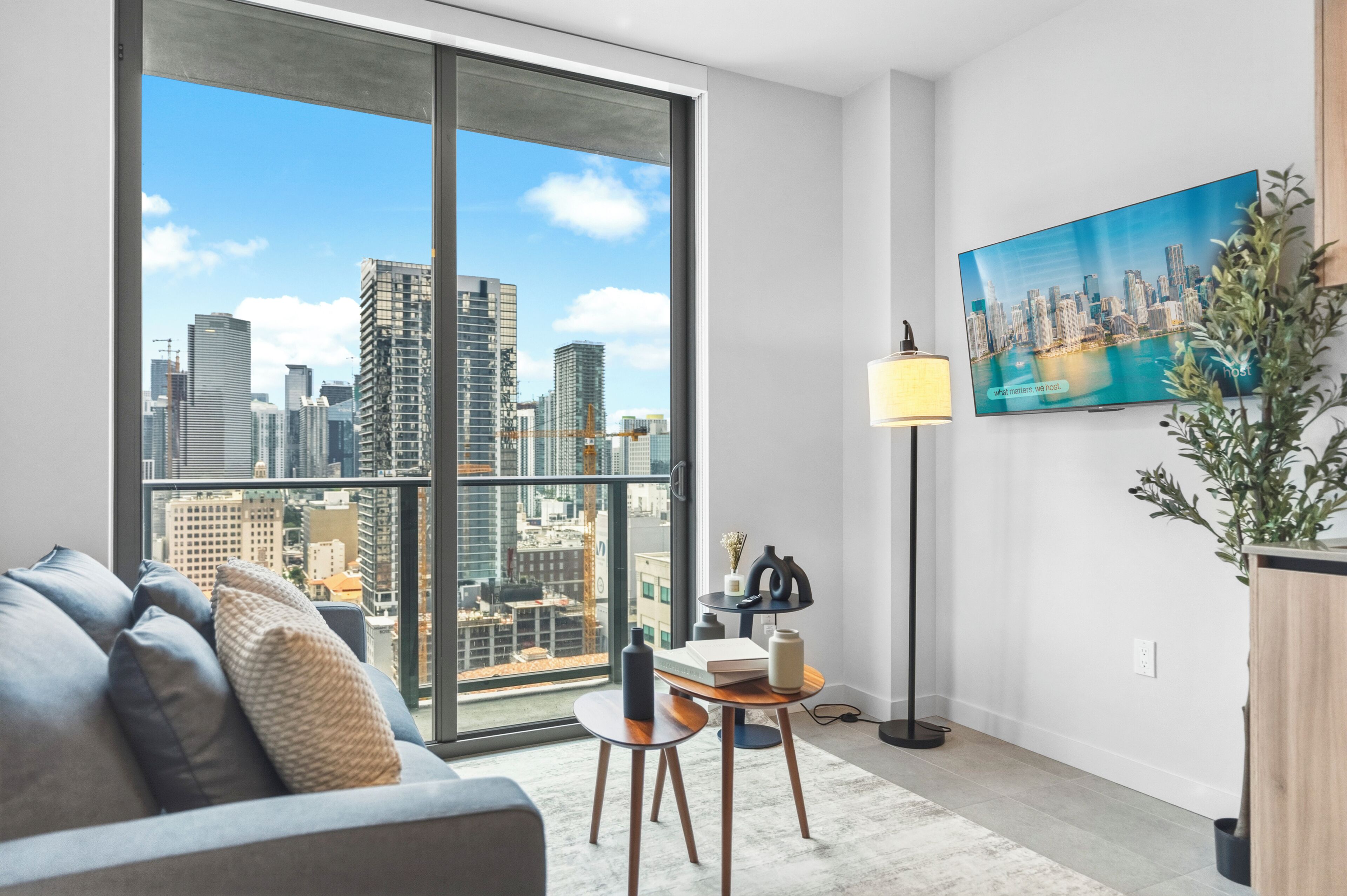 A modern living room with a large window, a sofa, a small table, a floor lamp, and a painting of a cityscape.