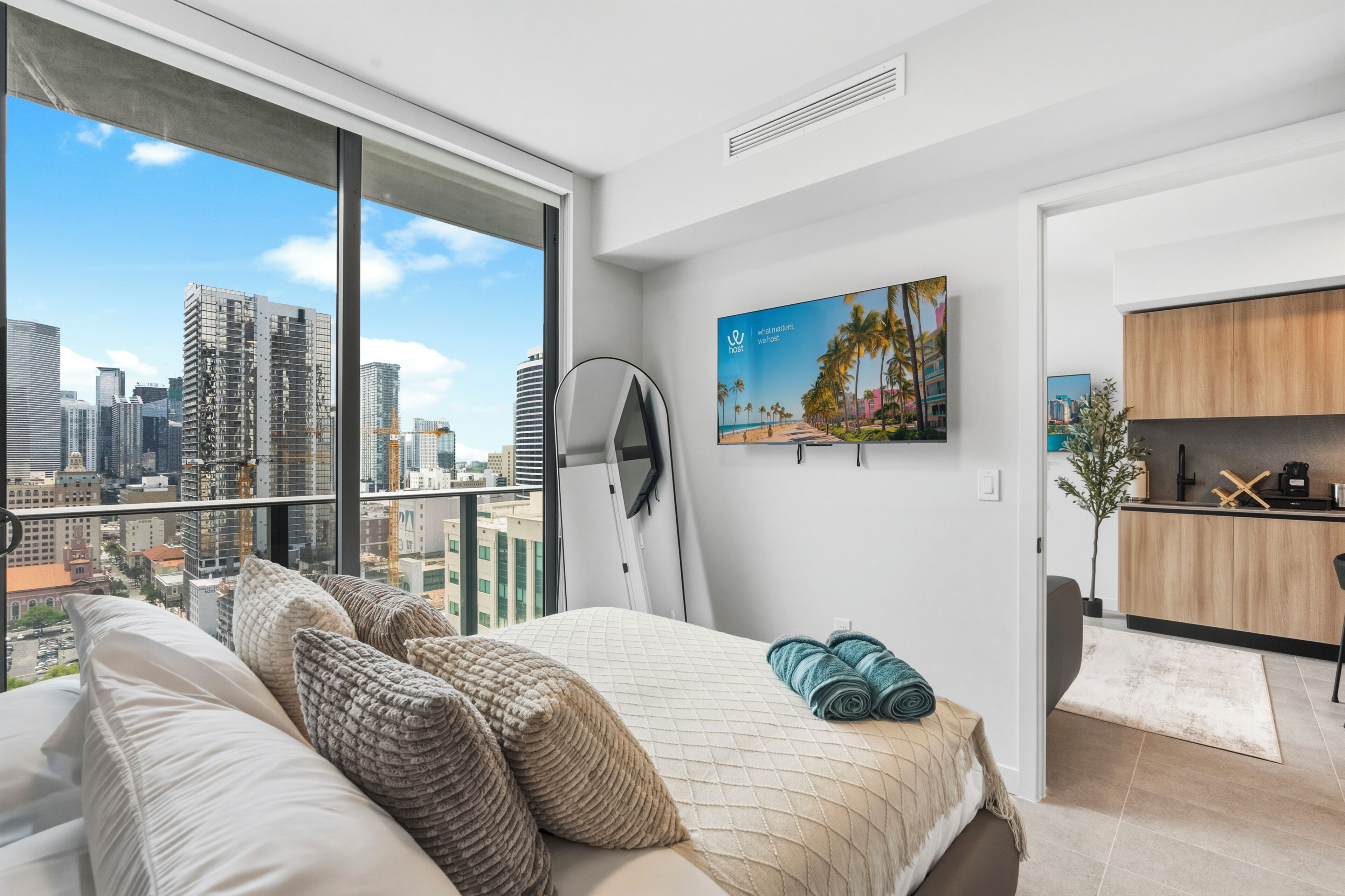 Habitación de hotel moderna con un ventanal grande, una cama con almohadas, un cuadro, una zona de cocina y vistas al horizonte de la ciudad.