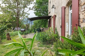 Un edificio rústico de piedra con un porche cubierto, una parrilla y abundante vegetación.