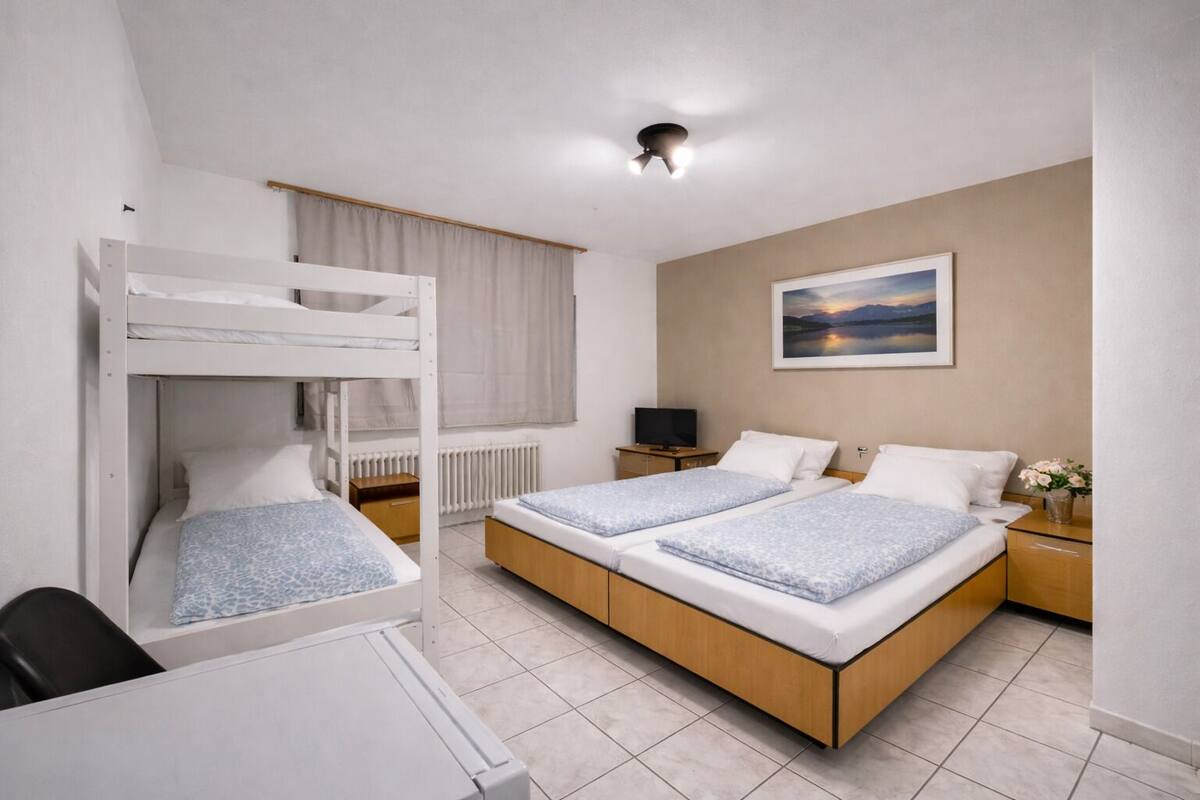 Um quarto com uma cama beliche, uma cama de casal, uma escrivaninha, uma cadeira, uma televisão, um criado-mudo com um vaso de flores e um quadro na parede.