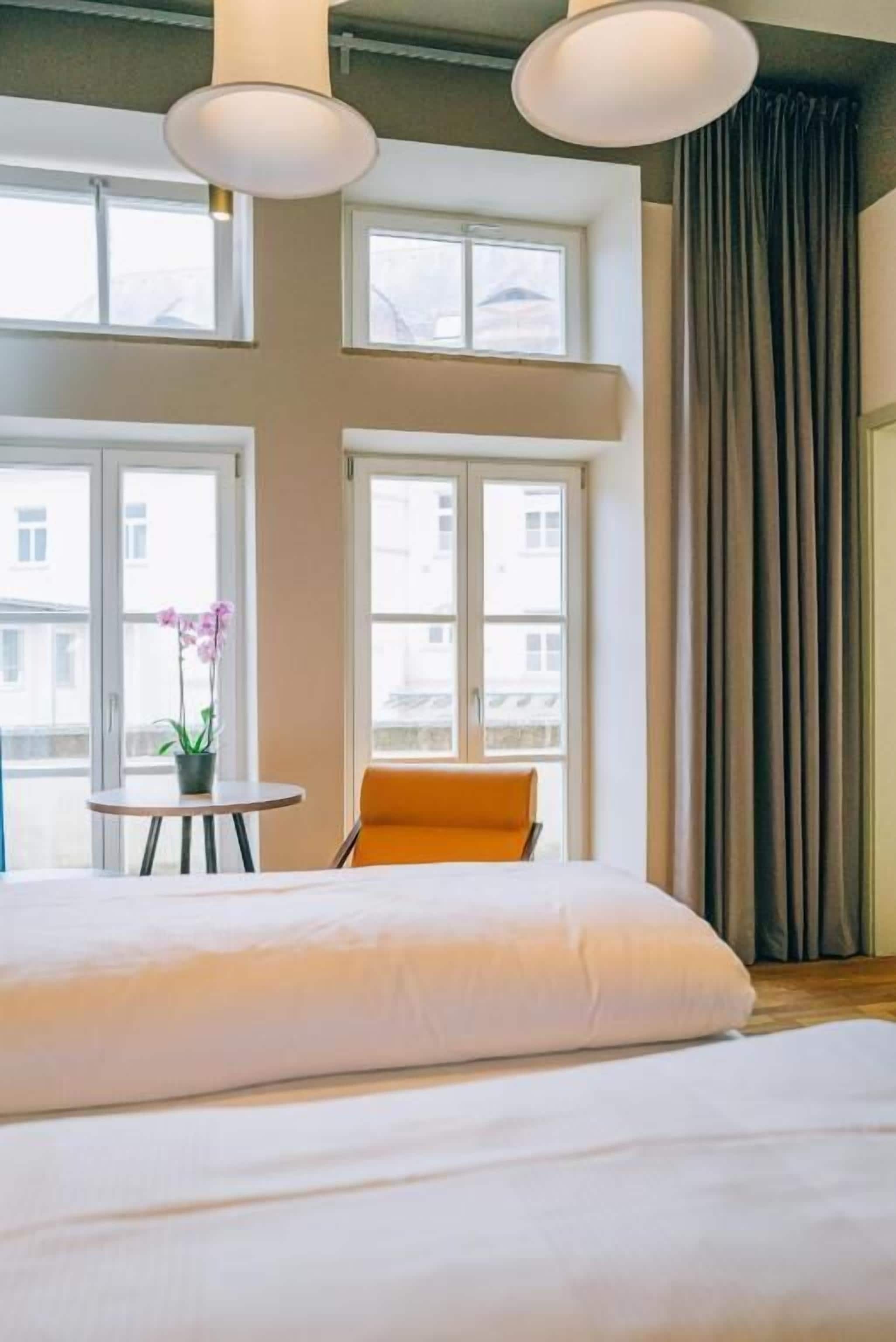 A hotel room with a large bed, two windows, a small table with a plant, and an orange chair.