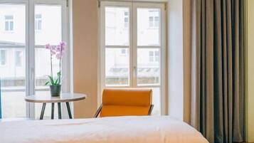 A hotel room with a large bed, two windows, a small table with a plant, and an orange chair.