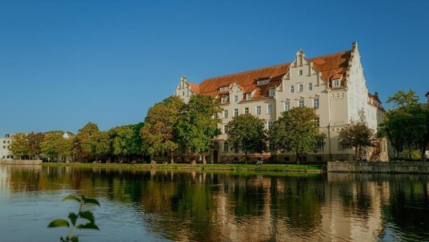 A large, historic building with multiple stories and a red-tiled roof, situated beside a calm body of water. - Hotel Amalia - Boutique Hotel (Landshut)