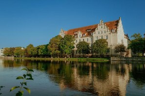 Ein großes, historisches Gebäude mit mehreren Etagen und einem roten Ziegeldach, neben einer ruhigen Wasserfläche gelegen.