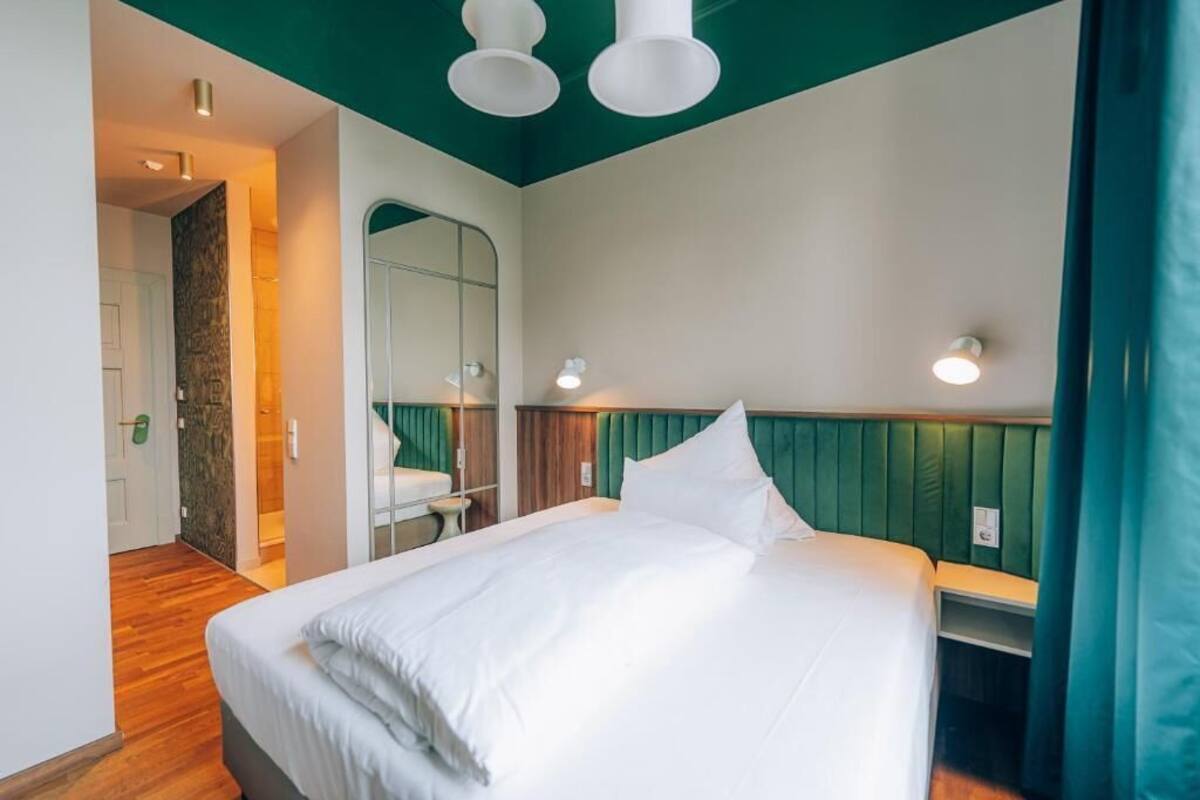 Una habitación de hotel moderna con una cama grande, piso de madera, una pared de acento verde y un espejo sobre la cama.