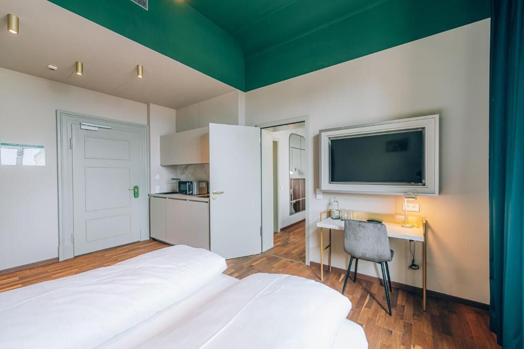 Quarto de hotel com duas camas, uma secretária com cadeira, televisão na parede e um espaço com cozinha compacta.
