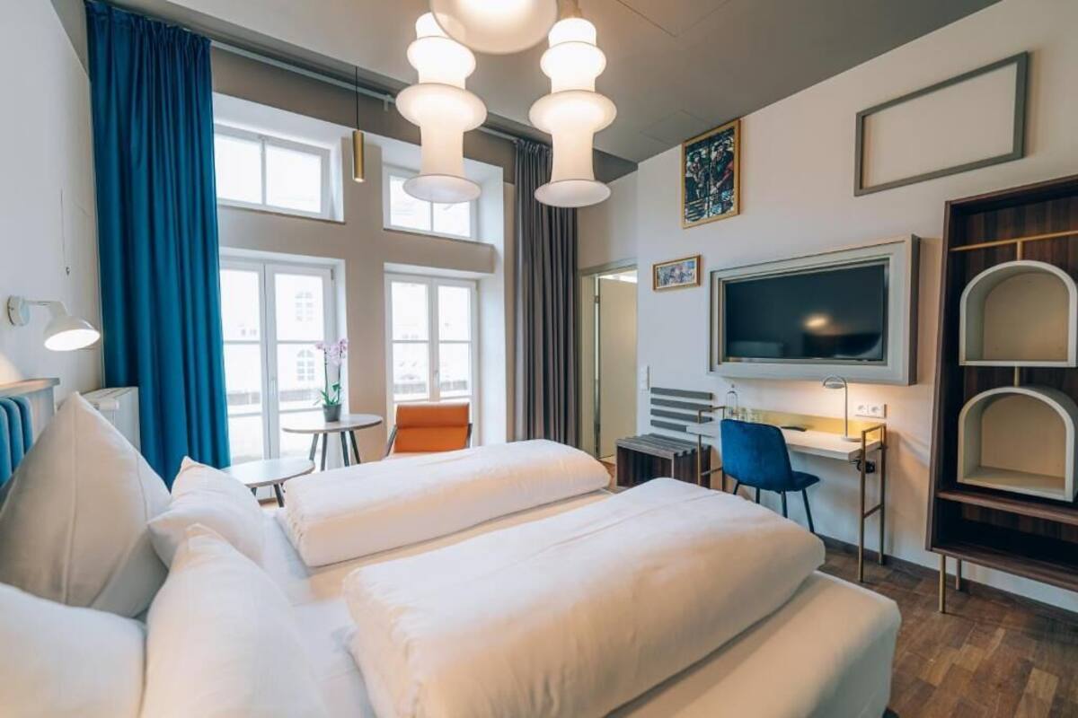 Habitación de hotel con cama, escritorio, silla, televisión y ventana con cortinas.
