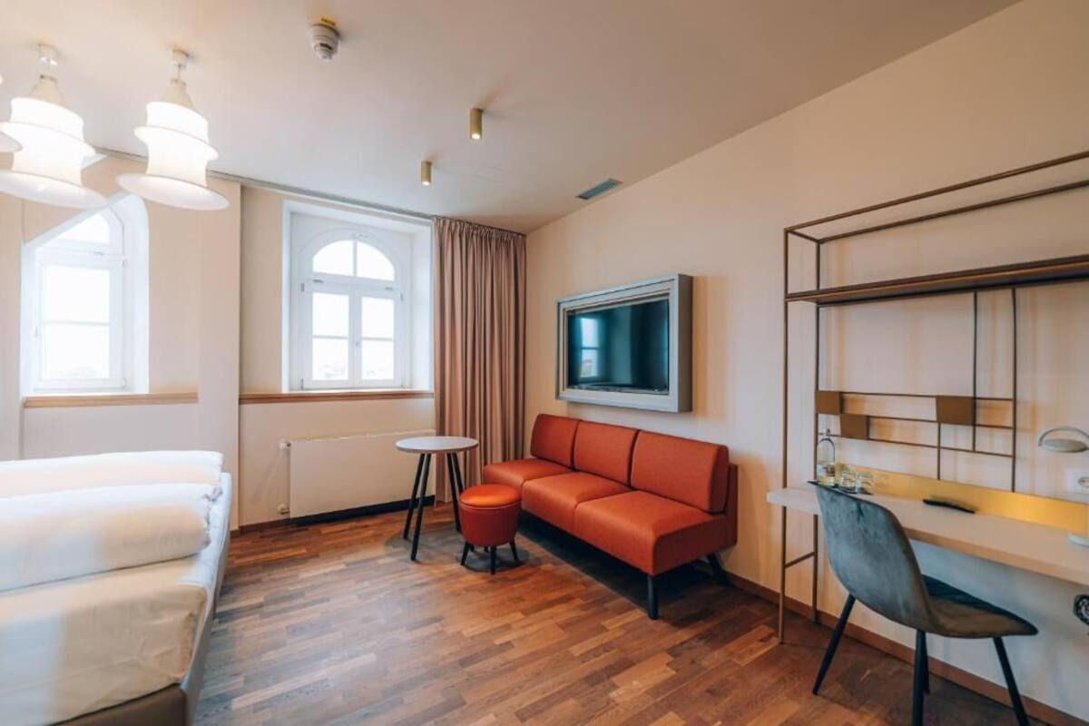 Una habitación de hotel moderna con suelo de madera, un sofá, un escritorio, una silla, un televisor y una ventana con cortinas.