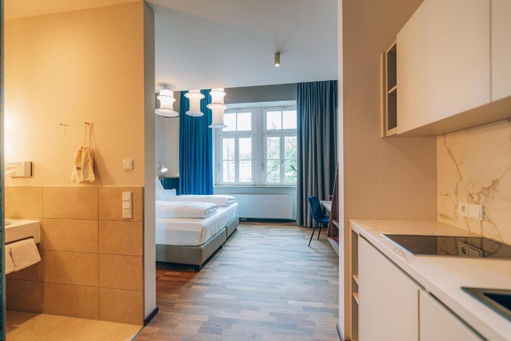 A modern hotel room with a bed, a small desk, a window with curtains, and a kitchenette.