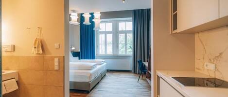 A modern hotel room with a bed, a small desk, a window with curtains, and a kitchenette.