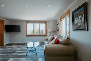 Una sala de estar con sofá, una mesa de centro de vidrio, un televisor montado en la pared y una ventana con vista a árboles.
