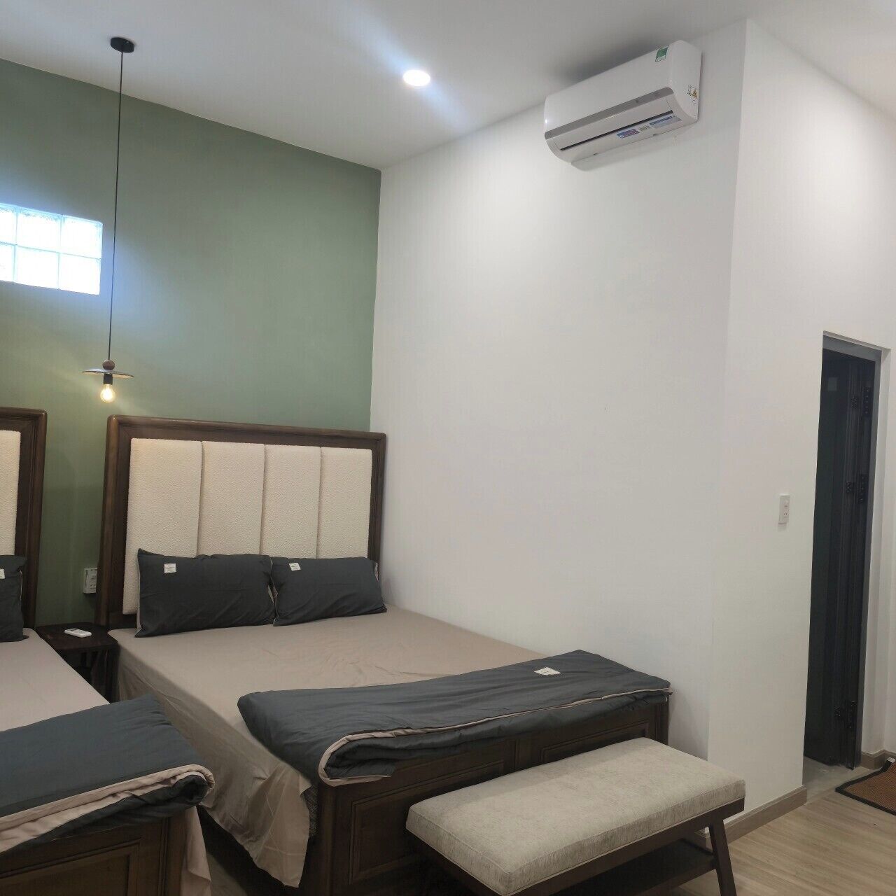A room with two beds, a bench, an air conditioning unit, a window, and a door.