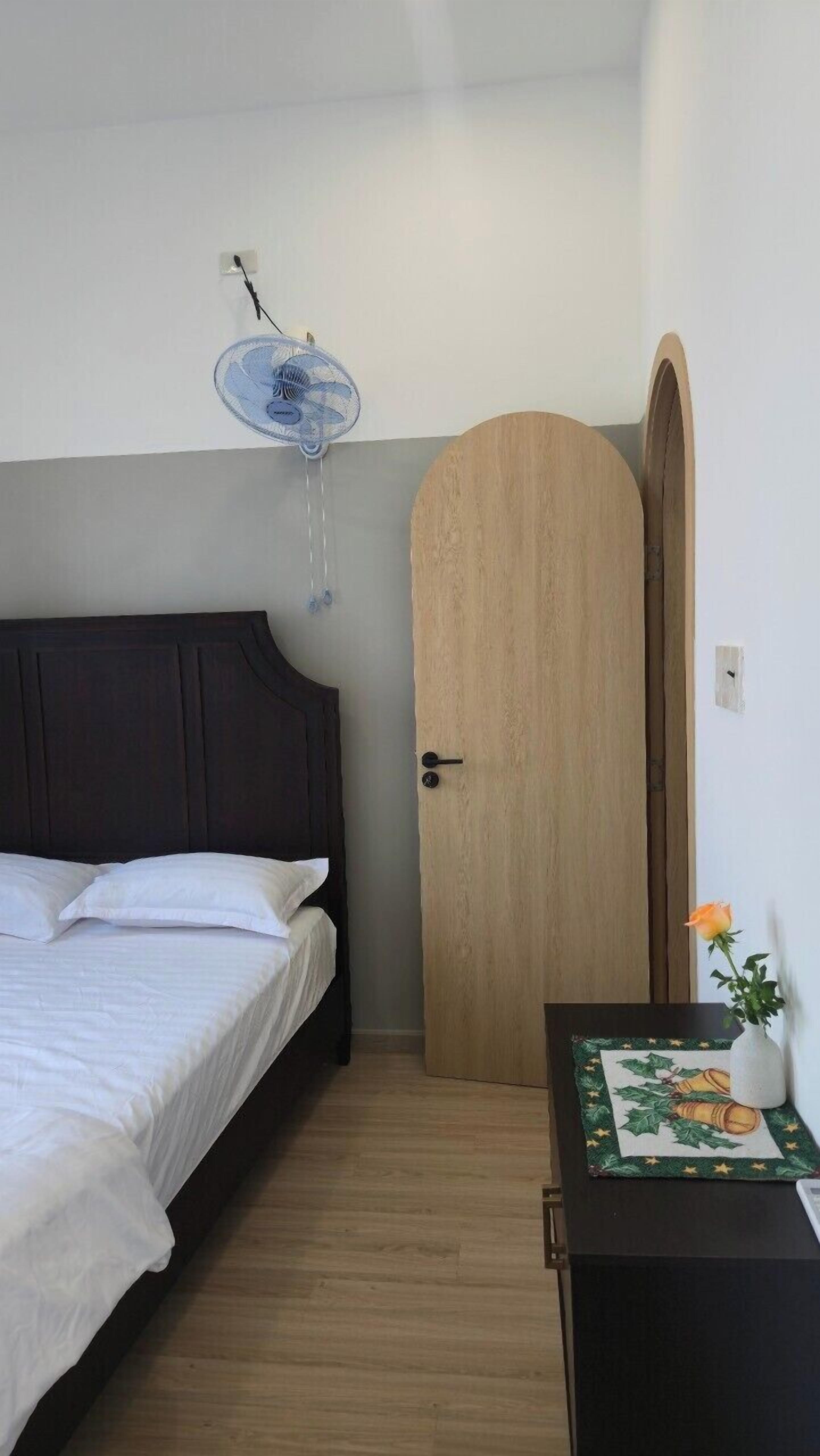 A bedroom with a bed, a wooden door, a small table with a vase, and a wall-mounted fan.