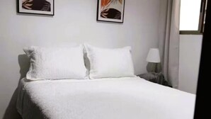 Una cama bien hecha con sábanas y almohadas blancas, una lámpara de noche y dos cuadros en la pared.