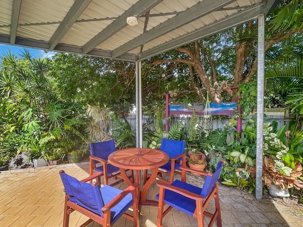Un área de estar exterior cubierta con una mesa de madera redonda y sillas azules, rodeada de abundante vegetación y plantas en macetas.
