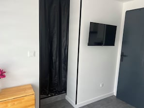 Quarto com um banco de madeira, cortina preta, TV de tela plana na parede e porta escura.
