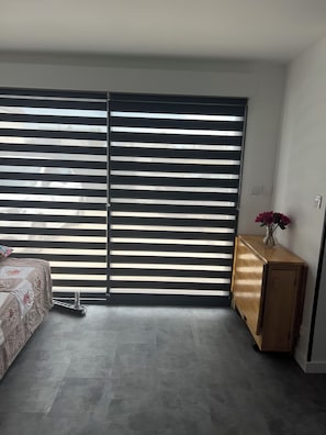 A bedroom with a bed, a bedside table with a vase, and a sliding door with horizontal blinds. - Erdivines – Ma Bro, La Suite, newly renovated studio, sleeps 2–4 – Erdeven (Erdeven)