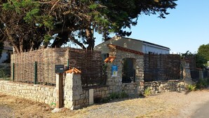 Un bâtiment en pierre avec un toit en tuiles, une enseigne indiquant « Chambres à louer », et un mur en pierre surmonté d’un portail en métal.