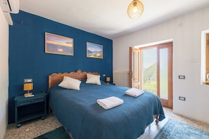 Une chambre avec un grand lit, deux tableaux accrochés au mur, une table de chevet avec une lampe et une porte donnant sur un balcon.