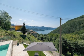 Un espace piscine agrémenté d’un grand parasol, avec vue sur un lac et sur une colline où se trouvent des maisons.