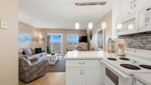 Een moderne keuken met witte kasten, een ingebouwde oven en een groot kookeiland. Er is een eethoek met een tafel en stoelen, en een woonkamer met een bank en een schilderij aan de muur.
