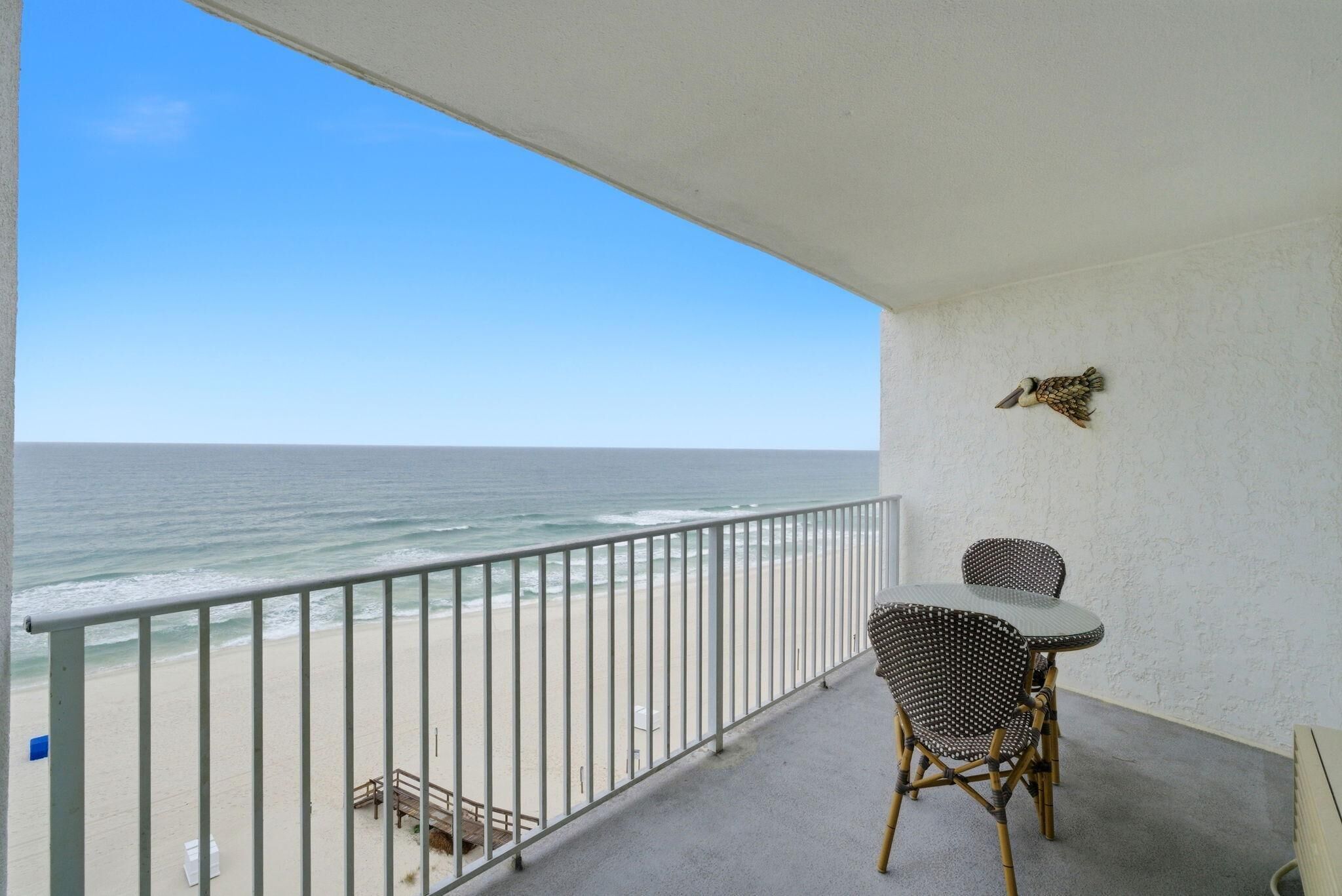 A balcony with a view of the beach and ocean. There are two wicker chairs and a small table.