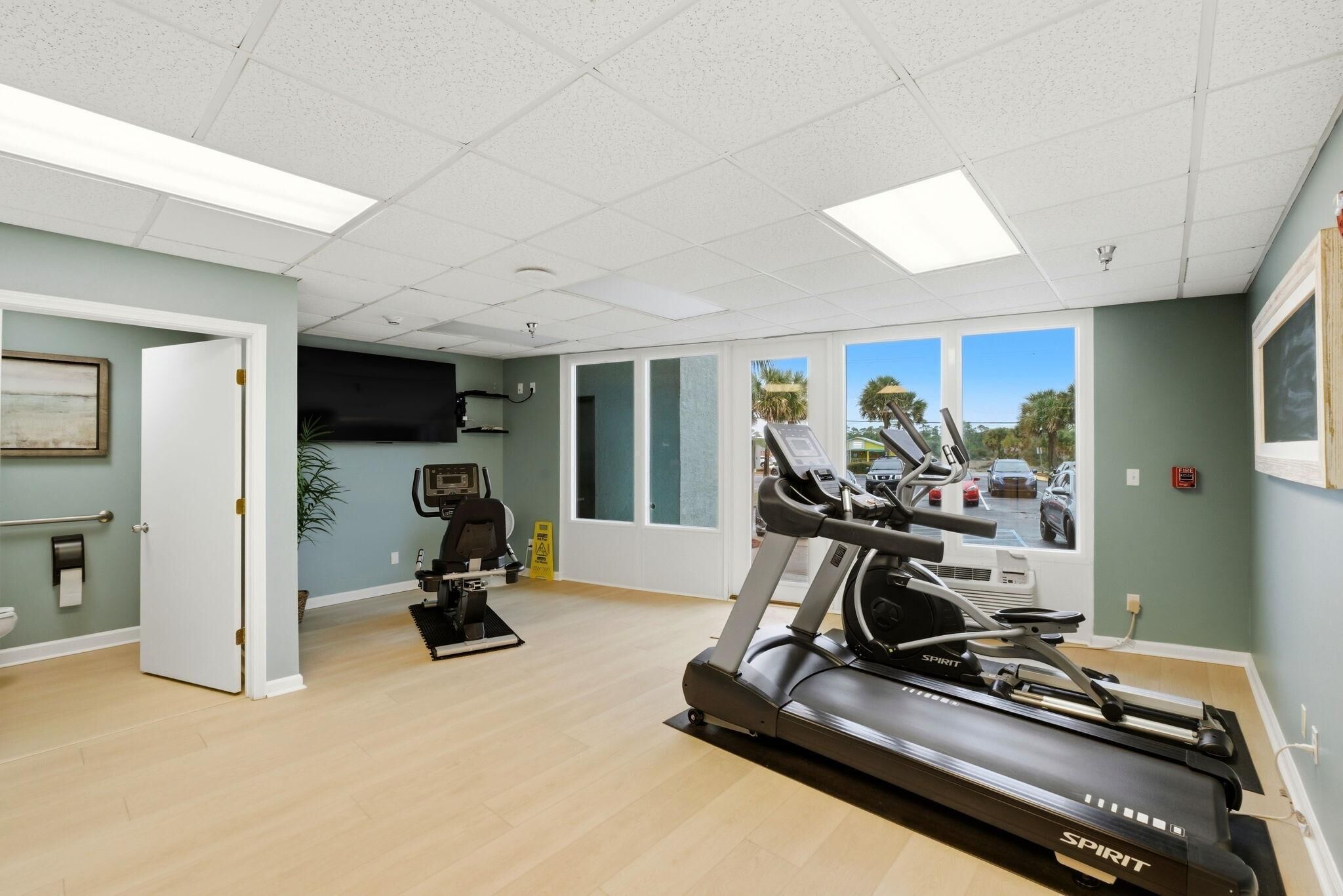 A gym with a treadmill, exercise bike, and a large window with a view of palm trees and cars.