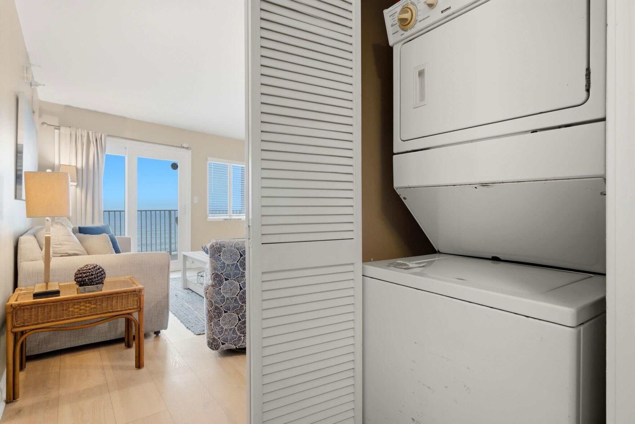 Ein Waschraum mit einer weißen Waschmaschine und einem Trockner, ein Korbtisch mit Lampe sowie ein Blick auf einen Balkon mit Sofa und Vorhängen.
