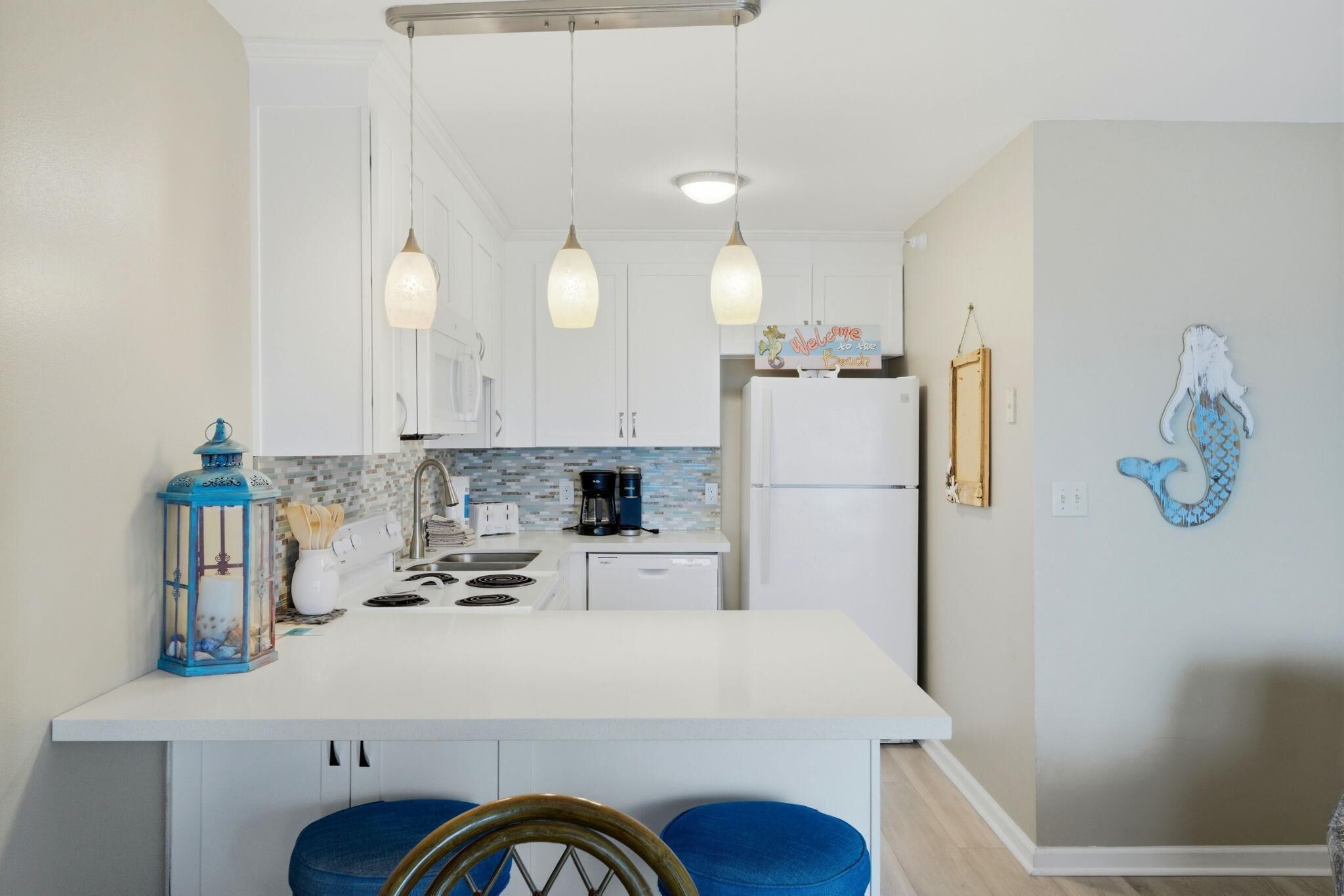 Une cuisine moderne avec des meubles blancs, un îlot central doté de tabourets, un éclairage suspendu et des œuvres d’art décoratives sur les murs.