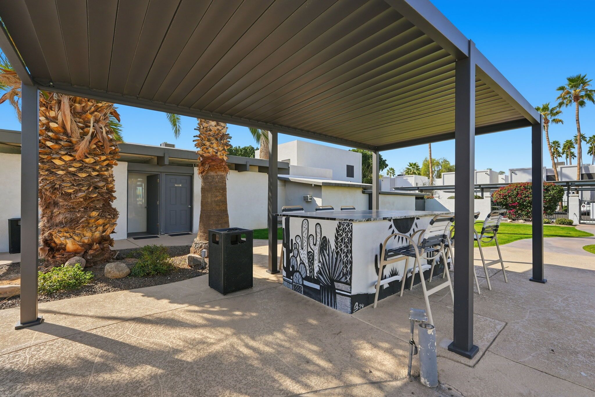 En overdækket patio med grill, udendørs møbler, palmer og en moderne bygning i baggrunden.