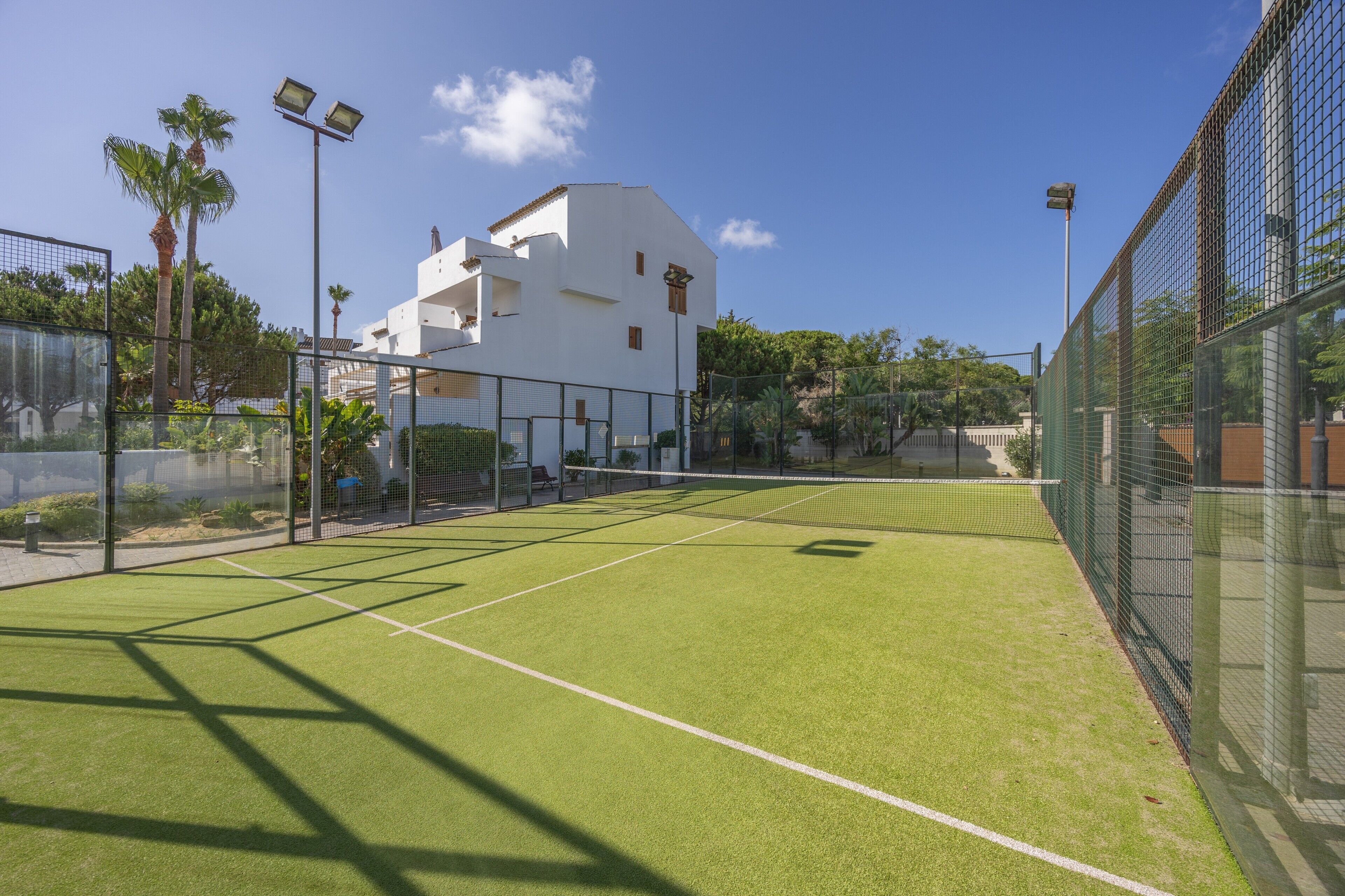 Una cancha de tenis al aire libre con red, rodeada por una cerca, y un edificio blanco al fondo.