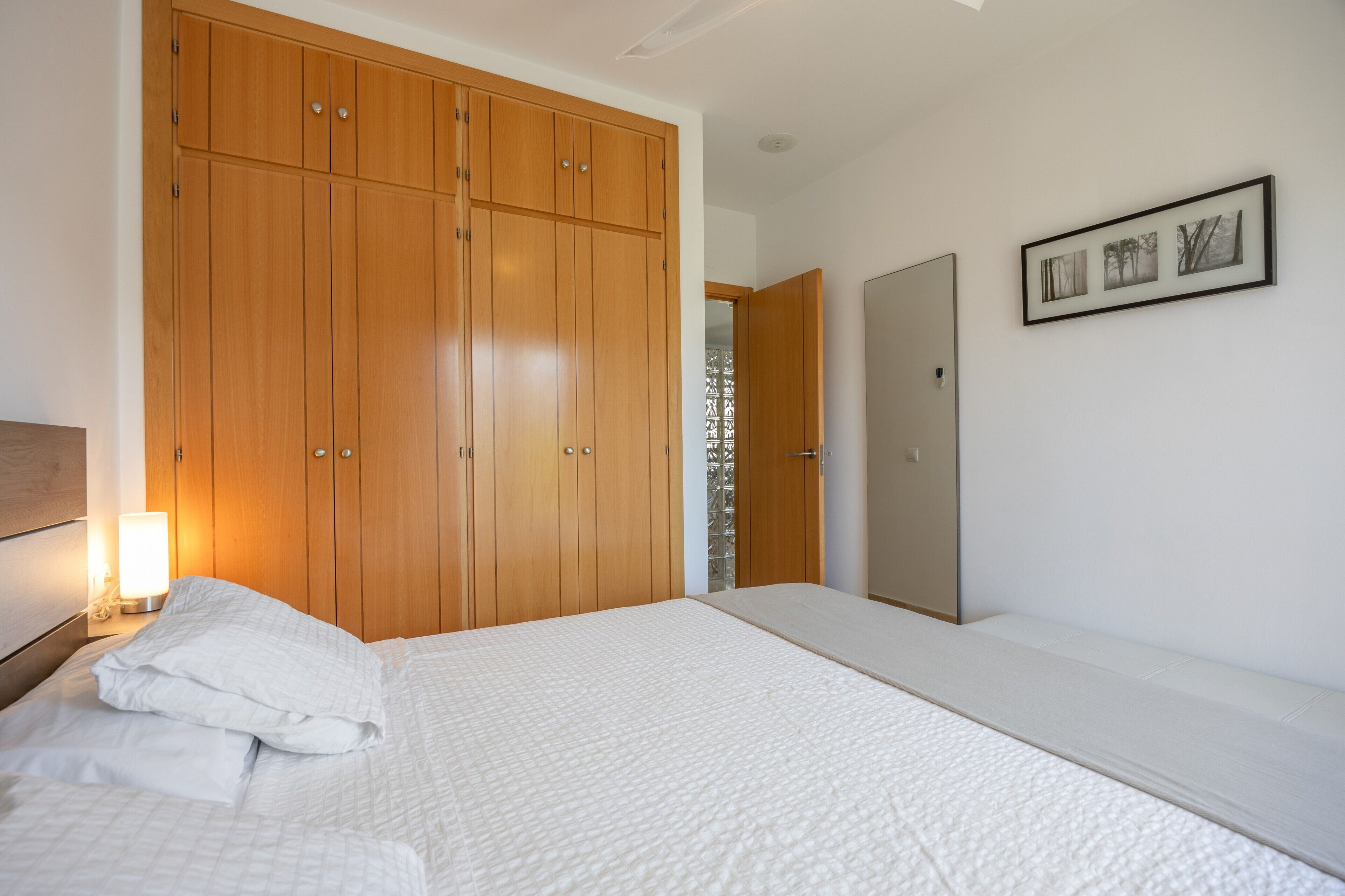 Ein Schlafzimmer mit großem Kleiderschrank, einem Bett mit weißer Bettwäsche, einer Nachttischlampe und einem gerahmten Bild an der Wand.
