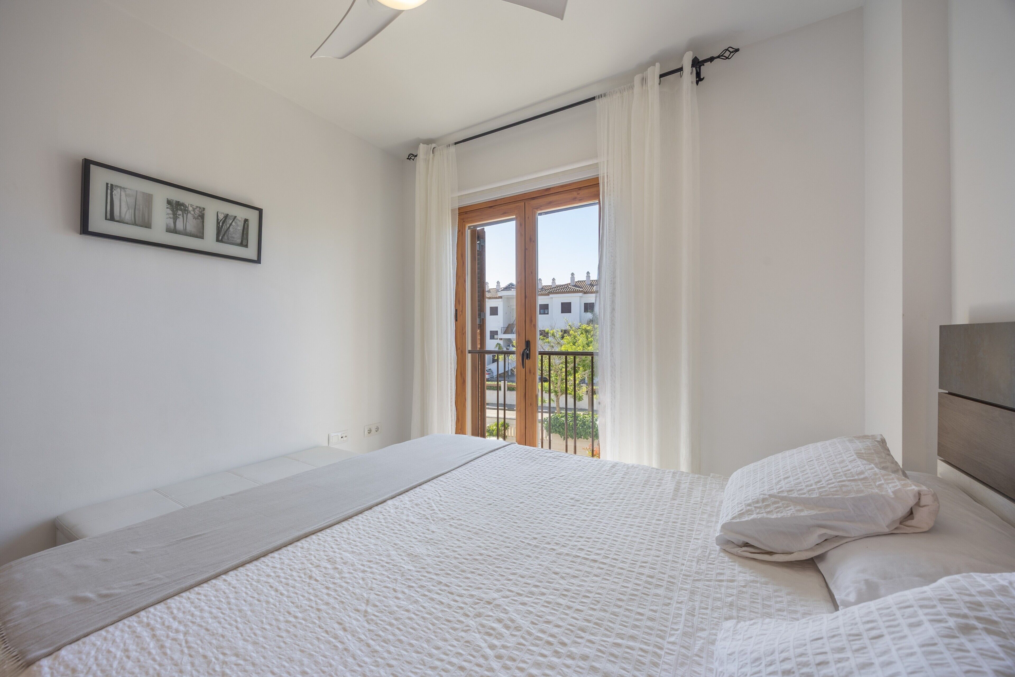 Un dormitorio con cama, ventana con cortinas, vista a edificios y un cuadro en la pared.