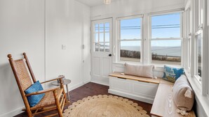 Una habitación con una mecedora de madera, un banco con cojines, un tapete redondo tejido y vistas al océano a través de amplias ventanas.