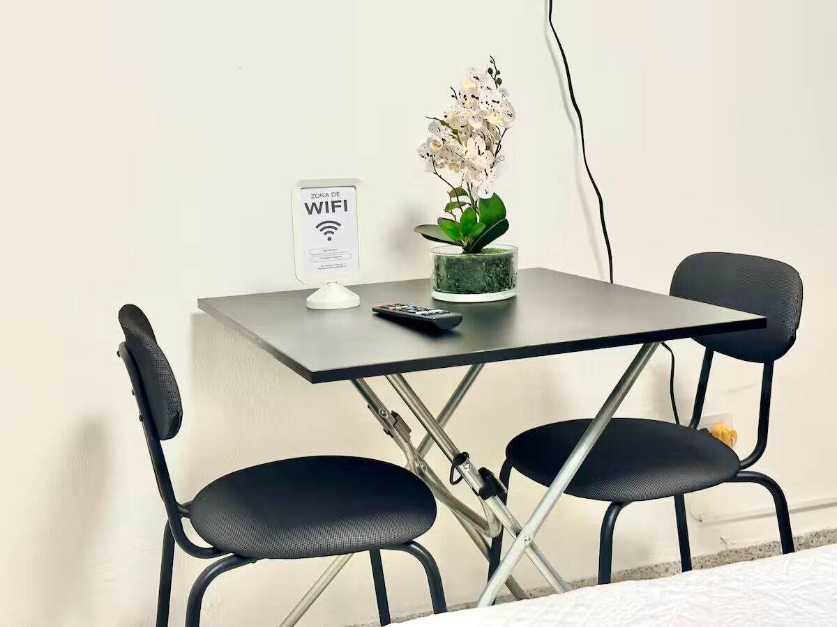 Et lille, moderne arbejdsområde med et klapbord, to sorte stole, en potteorkidé, en fjernbetjening og et WiFi-skilt på væggen.