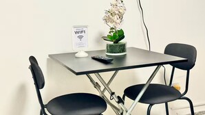 Un espacio de trabajo pequeño y moderno con una mesa plegable, dos sillas negras, una orquídea en maceta, un control remoto y un cartel de WiFi en la pared.