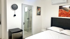 Una habitación de hotel moderna con cama, mesita de noche, reloj, cabina de ducha de vidrio, un pequeño calentador y un equipo de aire acondicionado.