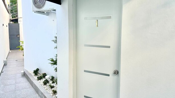 Una puerta blanca con una placa de identificación, un equipo de aire acondicionado pequeño sujeto a la pared, y un sendero estrecho bordeado de plantas en macetas.